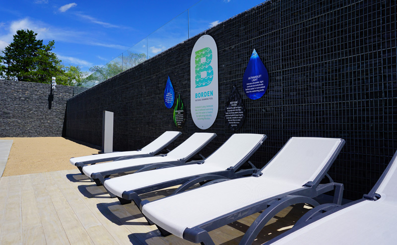Beach chairs lined up on the edge of Borden Natural Swimming Pool. A sign with the pool's logo is on a wall behind the chairs.
